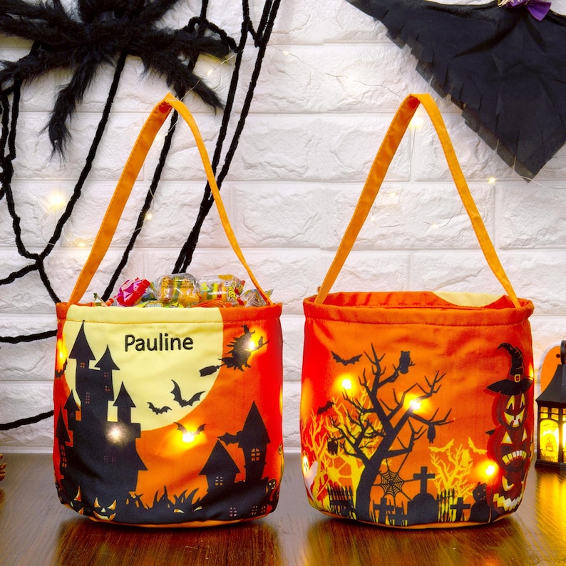 May include: Two orange Halloween treat bags with orange handles. One bag features a black silhouette of a haunted house, bats, and the name "Pauline". The other bag has a spooky tree, bats, and a jack-o'-lantern. Both bags are illuminated with small lights.