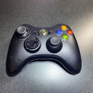 Puede incluir: Mando negro de Xbox 360 con dos palancas analógicas, una cruceta y botones de acción de colores. El mando tiene un acabado brillante y un logotipo plateado de Xbox en el centro.