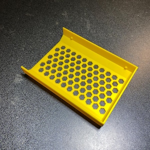 Puede incluir: Una bandeja rectangular de color amarillo brillante con un patrón de panal de agujeros hexagonales. La bandeja tiene bordes elevados en dos lados y está hecha de un material sólido, posiblemente plástico. El diseño sugiere un propósito funcional u organizativo.