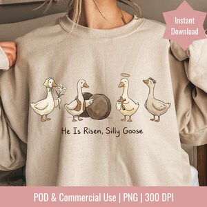 May include: Beige sweatshirt featuring a whimsical illustration of four geese. The geese are depicted with various accessories, including a bonnet, vest, halo, and floral crown. The text "He Is Risen, Silly Goose" is printed below the geese.