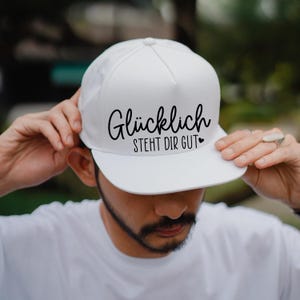 K&ouml;nnte beinhalten: Wei&szlig;e Baseballkappe mit schwarzem Text, der "Gl&uuml;cklich steht dir gut" mit einem kleinen Herzsymbol lautet.