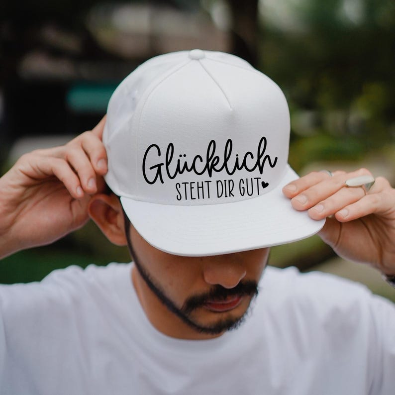K&ouml;nnte beinhalten: Wei&szlig;e Baseballkappe mit schwarzem Text, der "Gl&uuml;cklich steht dir gut" mit einem kleinen Herzsymbol lautet.