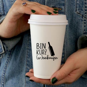 K&ouml;nnte beinhalten: Wei&szlig;er Coffee-to-go-Becher mit schwarzem Text "Bin kurz (w)einkaufen" und einer schwarzen Silhouette einer Weinflasche.