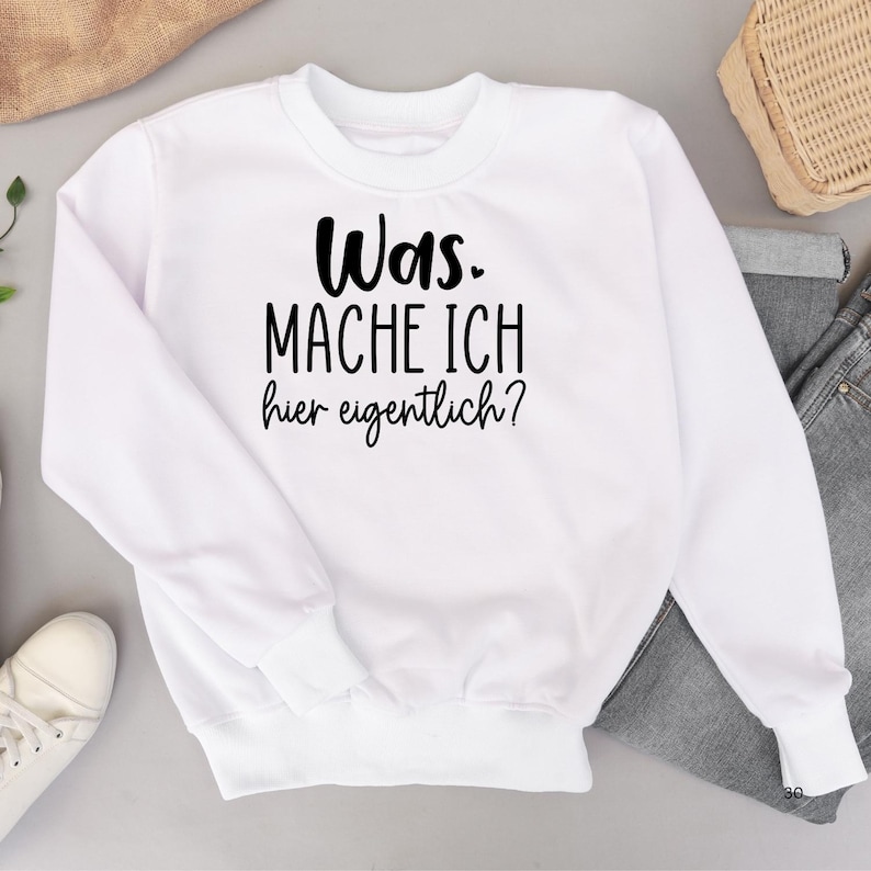 K&ouml;nnte beinhalten: Wei&szlig;er Pullover mit schwarzem Text, der "Was mache ich hier eigentlich?" lautet.