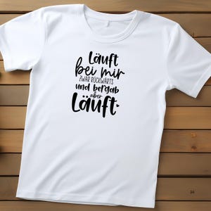 K&ouml;nnte beinhalten: Wei&szlig;es T-Shirt mit schwarzem Text, der "l&auml;uft bei mir zwar r&uuml;ckw&auml;rts und bergauf aber l&auml;uft." lautet.