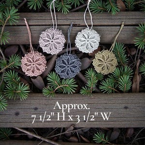 May include: Six macrame snowflake ornaments in various colors, including pink, gray, and beige, are displayed on a rustic wooden surface with pine needles. Each ornament is approximately 7.5 inches high and 3.5 inches wide, with a loop for hanging.