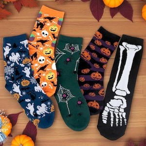 Puede incluir: Cinco pares de calcetines con temática de Halloween en varios colores. Los diseños incluyen fantasmas, calabazas, arañas y un esqueleto. Los calcetines están colocados sobre una superficie de madera con calabazas y hojas de otoño.
