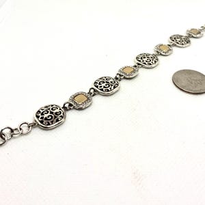 May include: A silver-tone link bracelet featuring alternating square filigree charms and smooth gold-tone square accents. The bracelet includes a lobster claw clasp and a small heart-shaped charm at the end, positioned next to a US quarter coin.