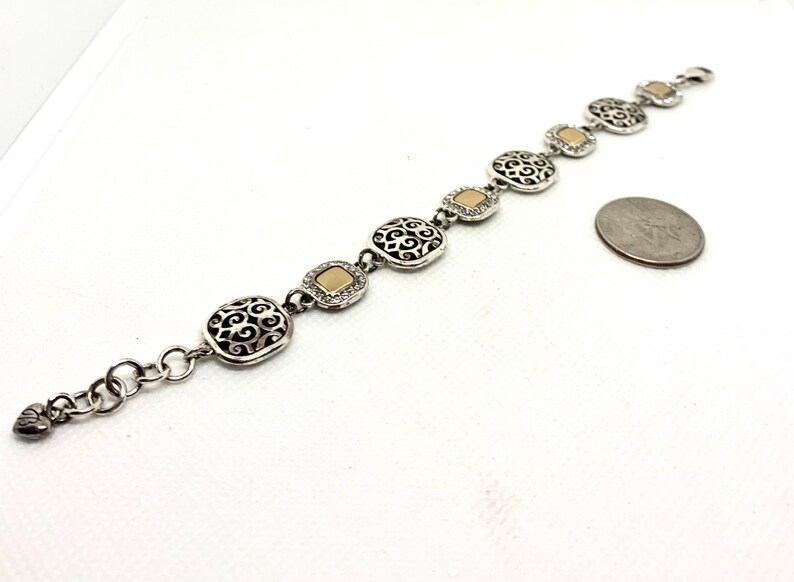 May include: A silver-tone link bracelet featuring alternating square filigree charms and smooth gold-tone square accents. The bracelet includes a lobster claw clasp and a small heart-shaped charm at the end, positioned next to a US quarter coin.