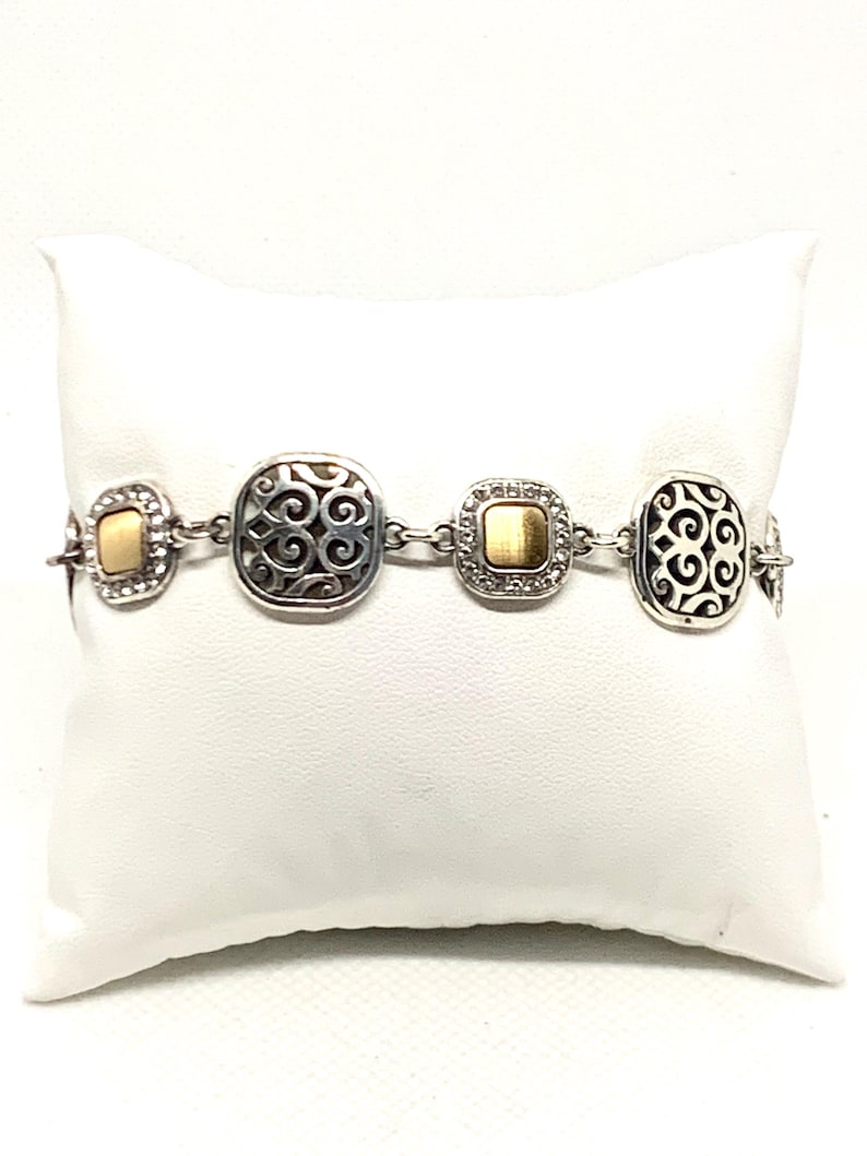 May include: A silver-tone link bracelet featuring alternating square and rectangular stations. Two stations display an openwork scroll pattern, while the other two feature a smooth, brushed gold-tone center framed by small, clear, sparkling stones.