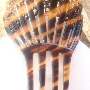 May include: An antique hair comb with a fan-shaped design. The comb features a tortoiseshell pattern with alternating bands of dark brown and amber. The top portion has intricate, openwork detailing. The comb's teeth are long and evenly spaced.
