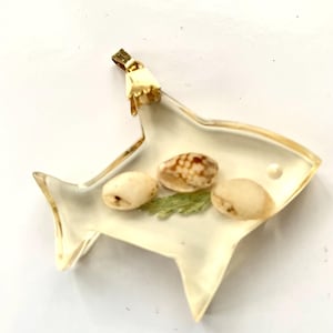 May include: A clear, fish-shaped pendant with gold-colored trim. The pendant contains three small, oval-shaped seashells and a sprig of green foliage. A gold-colored clasp is attached to the top of the pendant.