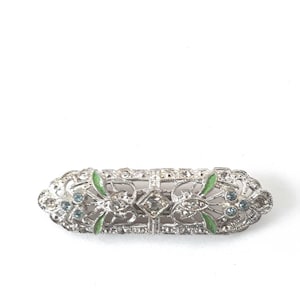 May include: An elongated silver-tone bar brooch featuring intricate filigree metalwork, small clear rhinestones, light blue accent stones, and vibrant green enamel leaf-shaped details arranged in a symmetrical, decorative pattern across the surface.