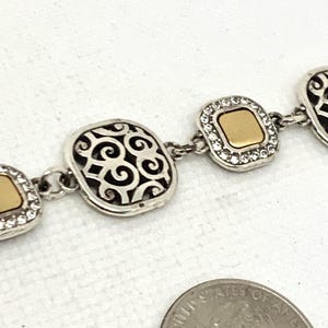 May include: A silver-tone link bracelet featuring alternating square-shaped charms. Some charms have a smooth gold-tone center surrounded by clear crystals, while others feature an openwork scroll pattern. A US quarter is shown for scale.