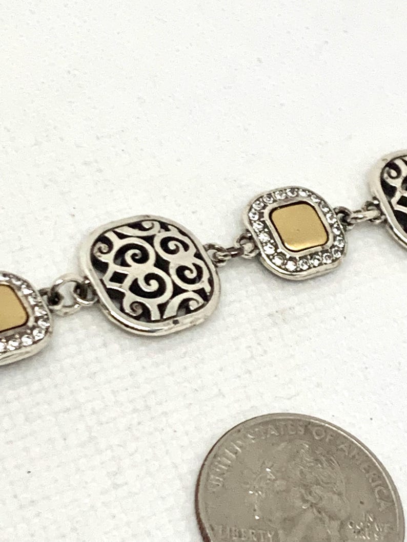 May include: A silver-tone link bracelet featuring alternating square-shaped charms. Some charms have a smooth gold-tone center surrounded by clear crystals, while others feature an openwork scroll pattern. A US quarter is shown for scale.