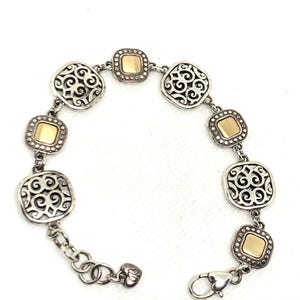 May include: A silver-tone link bracelet featuring alternating square charms. Three charms display an intricate openwork scroll pattern, while three charms feature a smooth, polished gold-tone center square bordered by a beaded silver-tone frame.