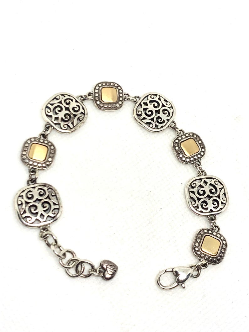 May include: A silver-tone link bracelet featuring alternating square charms. Three charms display an intricate openwork scroll pattern, while three charms feature a smooth, polished gold-tone center square bordered by a beaded silver-tone frame.