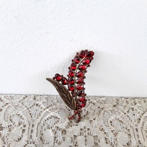 May include: A red and gold brooch in the shape of a stylized leaf with red rhinestones.