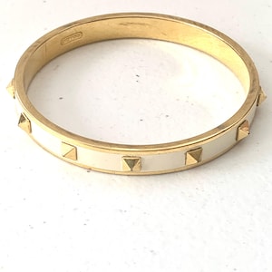 May include: Gold-tone metal bracelet with a cream-colored enamel band. The bracelet features gold-colored pyramid studs evenly spaced around the band. The Coach logo is visible on the inside of the bracelet.