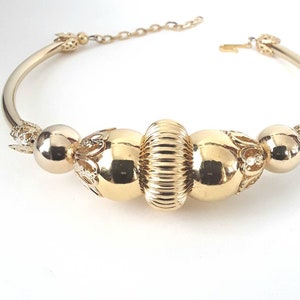 May include: A gold bangle bracelet with a decorative design featuring three large, round beads. The beads are intricately detailed with a textured surface and a floral pattern.
