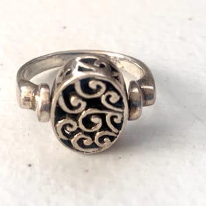 May include: Silver ring with an oval, openwork design. The center features a black background with swirling silver patterns. The ring has a thick band and decorative accents on the sides, showcasing a vintage aesthetic.