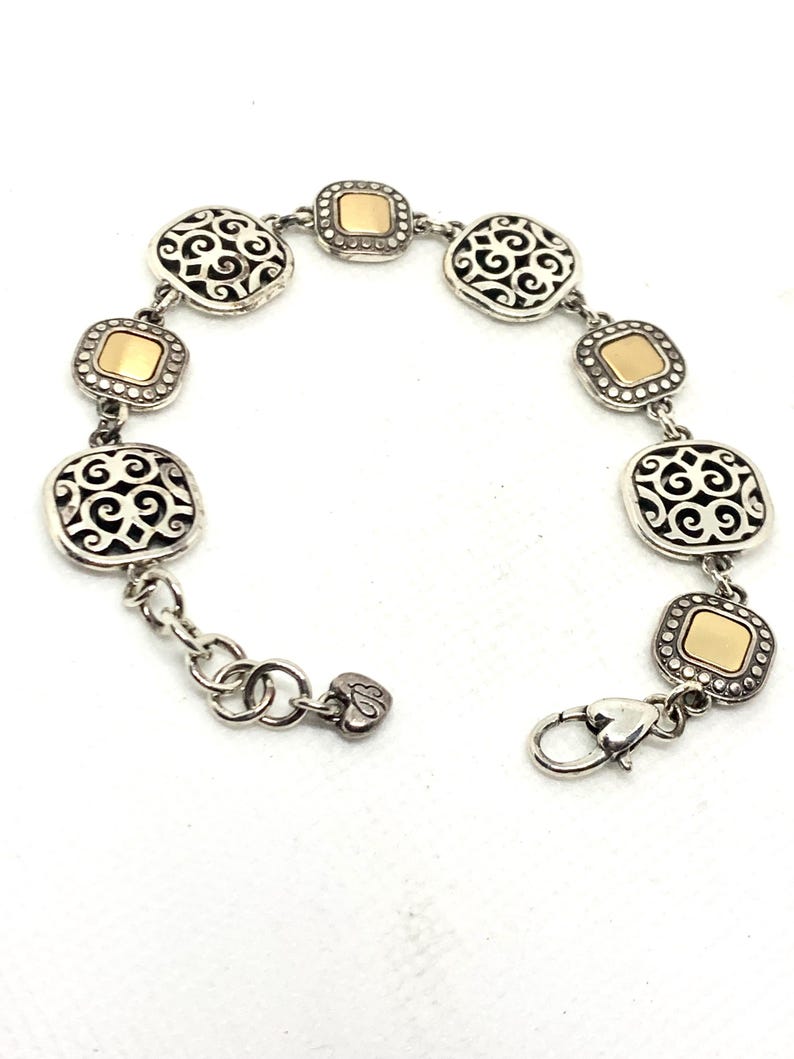 May include: A link bracelet featuring alternating square charms. Four charms display an intricate silver filigree scrollwork pattern, while three charms feature a smooth, polished gold-tone center framed by a silver beaded border. A heart-shaped charm hangs near the lobster claw clasp.