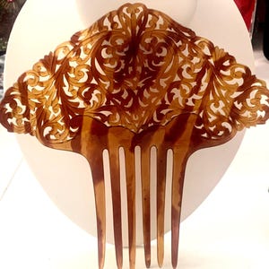 May include: A decorative hair comb with a fan-shaped top and long teeth. The comb is a warm brown color with intricate cut-out floral designs. The comb is displayed against a white background.