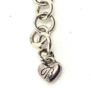 May include: A silver-toned metal chain extension featuring a series of circular links and a small heart-shaped charm at the end. The heart charm is engraved with a stylized letter B in a cursive font, providing a decorative finish to the jewelry piece.