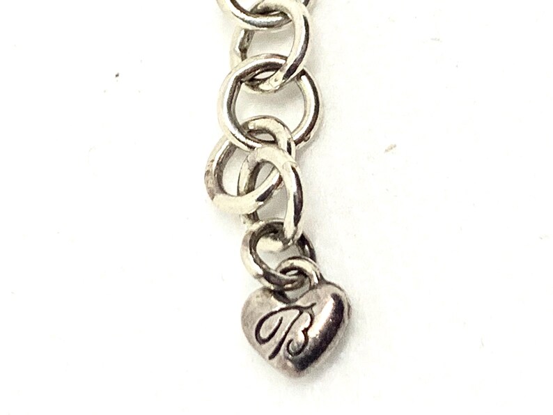 May include: A silver-toned metal chain extension featuring a series of circular links and a small heart-shaped charm at the end. The heart charm is engraved with a stylized letter B in a cursive font, providing a decorative finish to the jewelry piece.