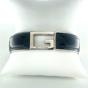 May include: Black leather bracelet with a silver-tone metal clasp. The clasp features the brand name "GUCCI" and a large "G" design. The bracelet is displayed on a white pillow.