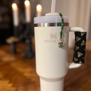 May include: A white Stanley tumbler with a handle wrapped in a black and gold pattern. A green beaded charm with a gold emblem hangs from the lid. The tumbler has a white lid and a clear straw.