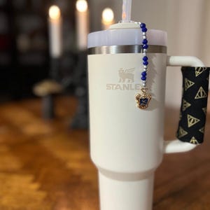 Puede incluir: Taza de viaje Stanley blanca con un charm de cuentas azules y plateadas con un escudo de la casa de Hogwarts dorado. La taza tiene un asa negra con un patrón dorado y negro.