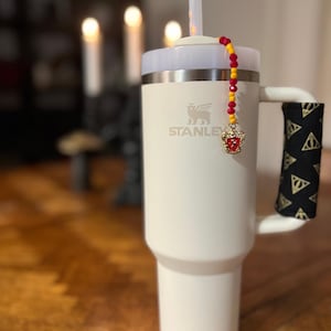 Puede incluir: Taza de viaje de acero inoxidable blanca con una envoltura de mango negra y dorada. La taza tiene un amuleto dorado con un escudo rojo y dorado que cuelga de una correa de cuentas rojas y amarillas.