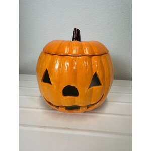 May include: A glossy orange ceramic pumpkin jar with a removable lid. The pumpkin features a carved jack-o'-lantern face with triangular eyes, a rounded nose, and a wide, curved mouth. A dark brown stem sits atop the lid of the round container.
