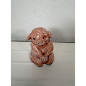 May include: A small, hand-sculpted ceramic pig figurine with a glossy, light brown finish and white painted brushstroke details. The pig has a rounded snout, small ears, and dark blue eyes, depicted in a seated position with its front paws together.