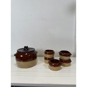 May include: A large ceramic pot with a lid and four smaller matching bowls, all featuring a two-tone design with a dark brown glazed upper section and a light tan lower section. Each piece has small side handles and a smooth, glossy finish.