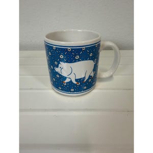 May include: A white ceramic mug featuring a blue background with a repeating pattern of small white flowers. A large, solid white pig illustration is centered on the front, with small orange accents on the pig's hooves. The mug has a white handle.