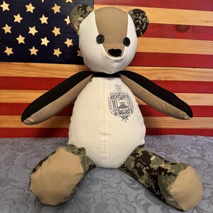 May include: A beige and camouflage teddy bear with black accents. The bear is wearing a white shirt with an embroidered crest that says "NAVY RESERVE." The bear is sitting in front of an American flag.