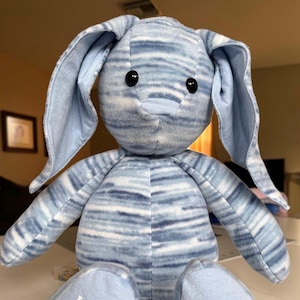 May include: A plush bunny toy with long, floppy ears and black button eyes. The bunny is made of soft fabric with a blue and white striped pattern. The toy is sitting on a white surface.