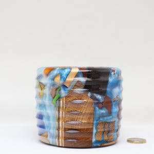 May include: A cylindrical wooden planter with a blue, white and brown abstract design. The planter has a ribbed texture and is made of wood and resin.