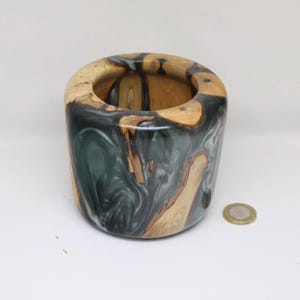 May include: A handcrafted wooden vase with a unique design. The vase features a combination of natural wood tones and a swirling, dark green and white resin. The cylindrical shape has a rounded top and a smooth finish. A coin is next to the vase.