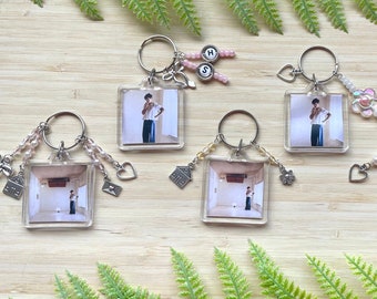 May include: Seven keychains with clear square photo frames featuring a person in each photo. Each keychain has a different charm attached, including a house, a flower, a heart, and a key. The keychains are on a wooden surface with green foliage.