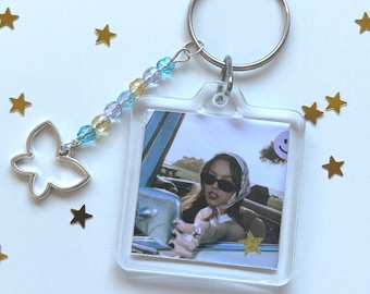 May include: A clear acrylic keychain with a photo of a woman wearing sunglasses and a headscarf driving a car. The keychain has a silver butterfly charm and a beaded chain with blue, yellow, and purple beads.