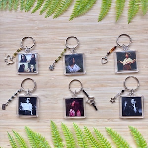 May include: Six clear acrylic keychains with silver rings and decorative charms. Each keychain features a different black and white or color photograph. Charms include a butterfly, flower, heart, and stars. The keychains are arranged on a wooden surface with green fern fronds.