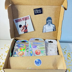 Könnte beinhalten: Ein brauner Karton mit verschiedenen Gegenständen. Darin befinden sich ein Shawn Mendes Albumcover, ein Ausschnitt einer Person und mehrere holografische Beutel mit Aufklebern. Die Box ist mit einem "Friends and Family"-Aufkleber und einem Discokugel-Aufkleber verziert.