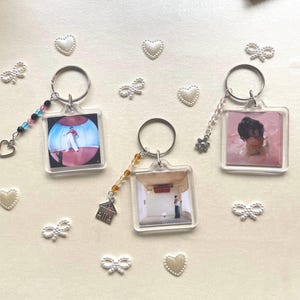 May include: Three clear plastic keyrings with different images inside. Each keyring has a silver ring and a decorative charm. The charms are a small house, a butterfly, and a string of colourful beads. The images are of a person in a white shirt and pink background, a person in a pink background, and a person in a white room.