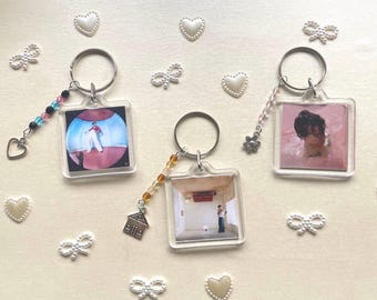 May include: Three clear plastic keychains with different images inside. Each keychain has a silver ring and a decorative charm. The charms are a small house, a butterfly, and a string of colorful beads. The images are of a person in a white shirt and pink background, a person in a pink background, and a person in a white room.