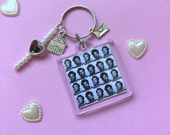 May include: A clear acrylic keychain featuring a grid of black and white portraits. The keychain has a silver ring with charms including a house, an envelope, and a heart-shaped key. The keychain is decorated with white pearl beads.