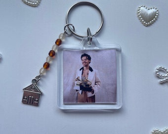 May include: Clear acrylic keychain featuring a square image of a person in a white suit. The keychain has a silver ring, a beaded strand with brown and white beads, and a small house charm. Decorative white hearts and bows surround the keychain.