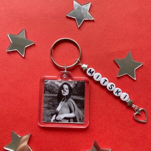May include: Clear square keychain with a black and white photo of a person. The keychain has a silver keyring and a beaded charm spelling "MITSKI". A silver heart charm and star charms are attached. The background is red with silver star decorations.